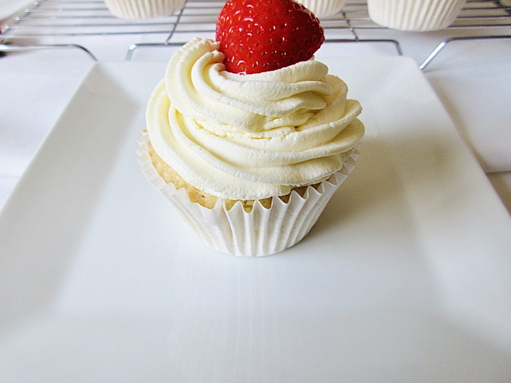 Single vanilla cupcake with whipped cream on top and half a strawberry on top of the whipped cream. The cupcake is on a square white small plate.