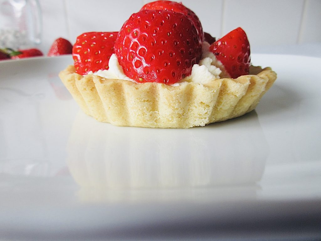 a single strawberry cream tartlet on a plate from a side view with white kitchen tiles as background.