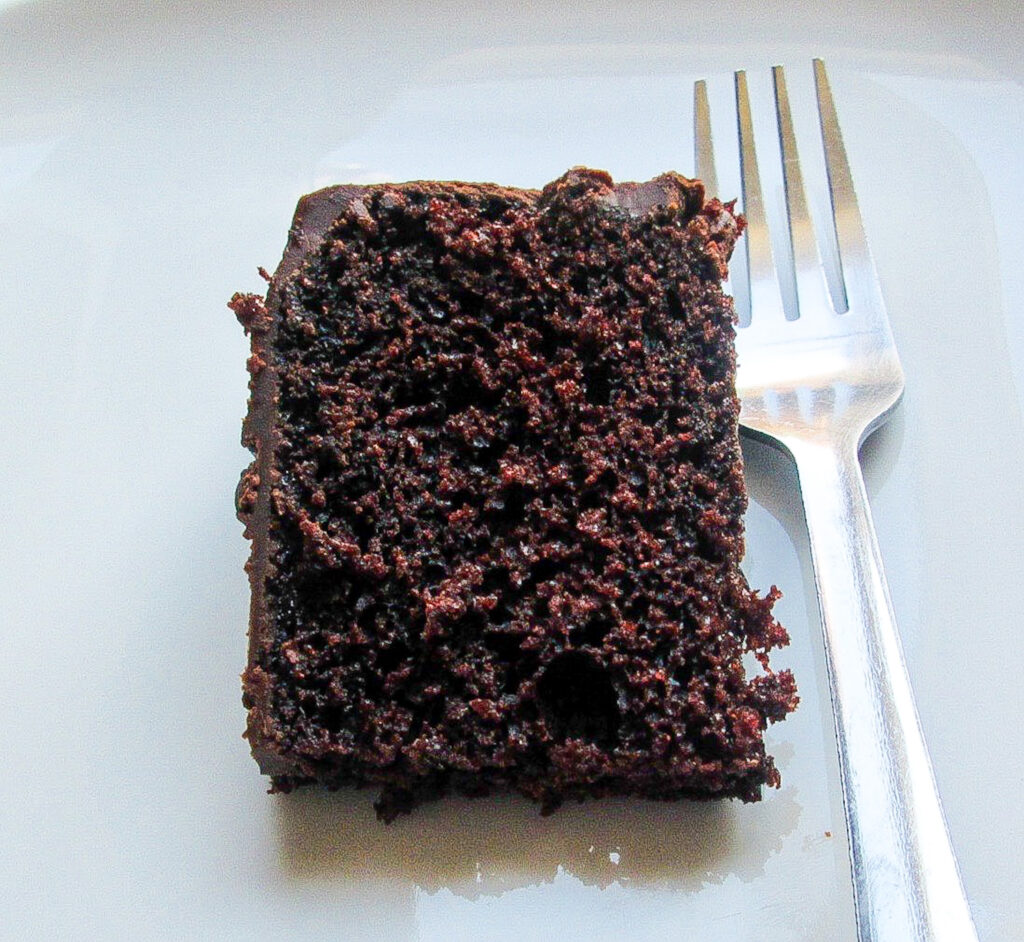 A slice of chocolate cake on a white plate with a silver fork next to the cake.