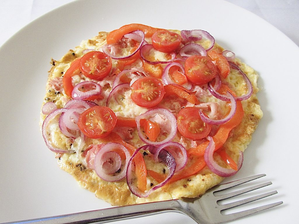 Omelette topped with cherry tomatoes, red onions and red peppers on a white plate with a fork next to it.