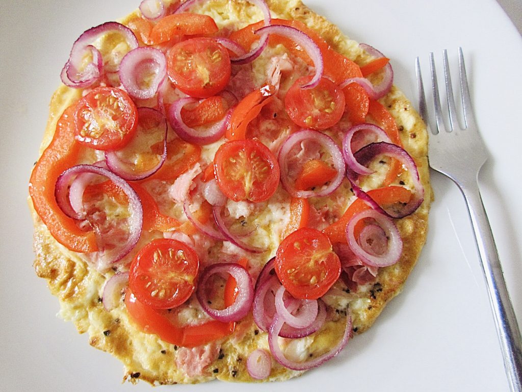 top view of omelette topped with cherry tomatoes, red onions and red peppers on a white plate with a fork next to it.