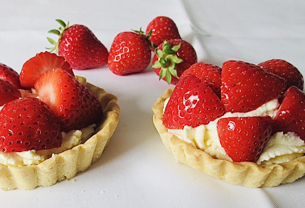 2 strawberry and cream tartlets on a white cloth with 4 strawberries behind them.