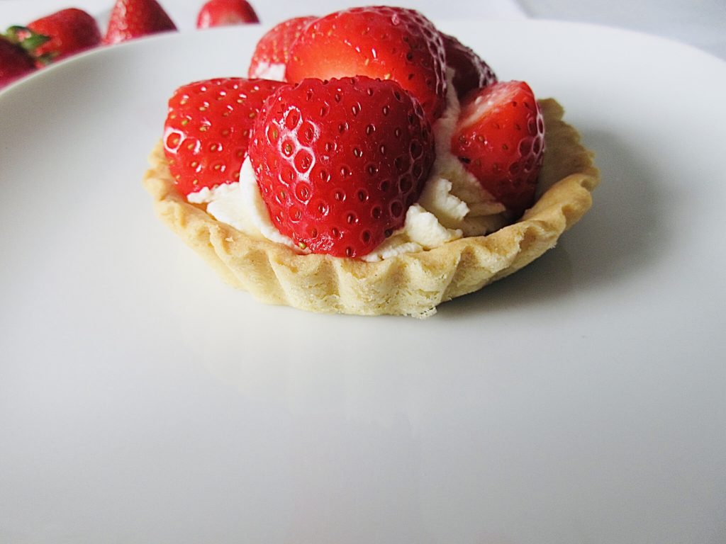 a single strawberry and cream mini tart on a white plate. 4 strawberries in the background.
