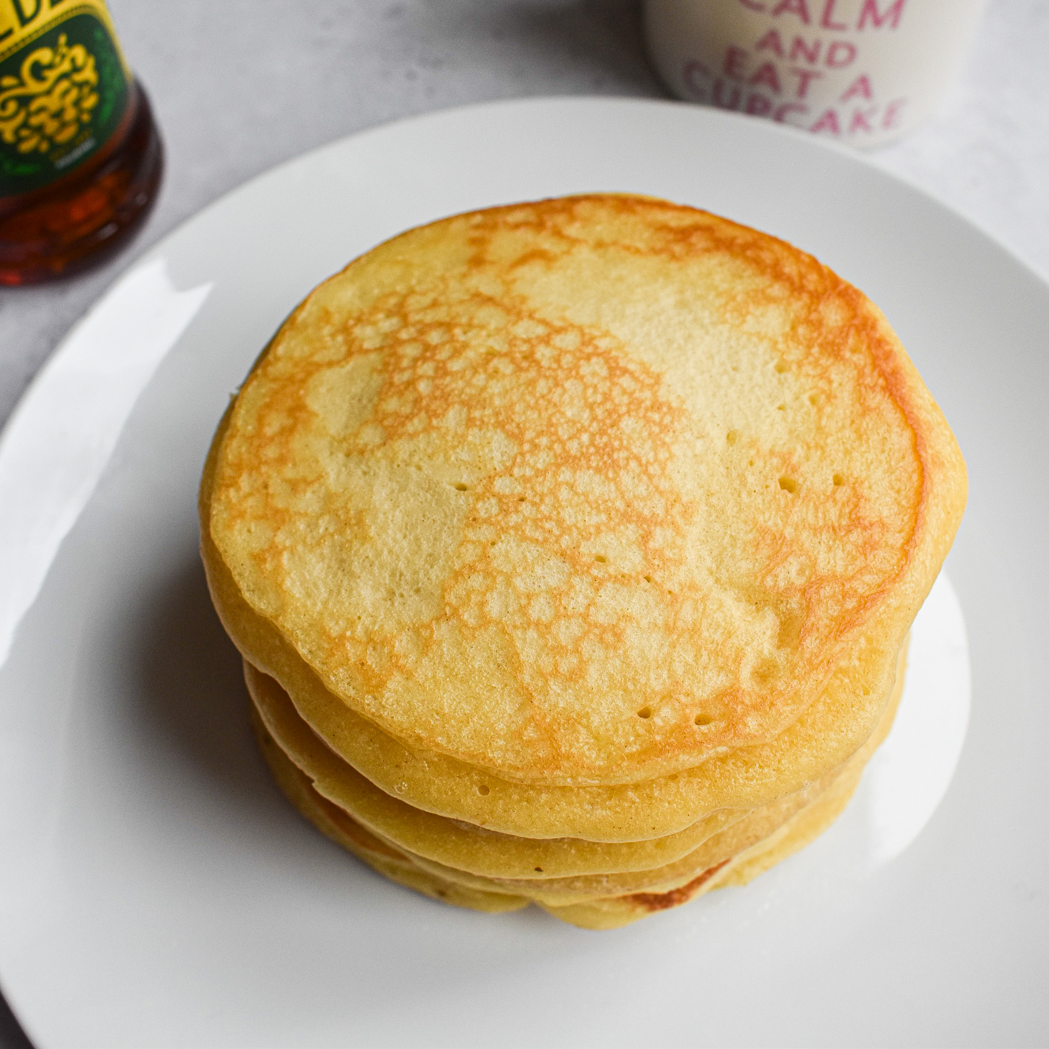 Pancakes stacked on a white plate. Part of a bottle of syrup in the background and a white mug with pink writing.