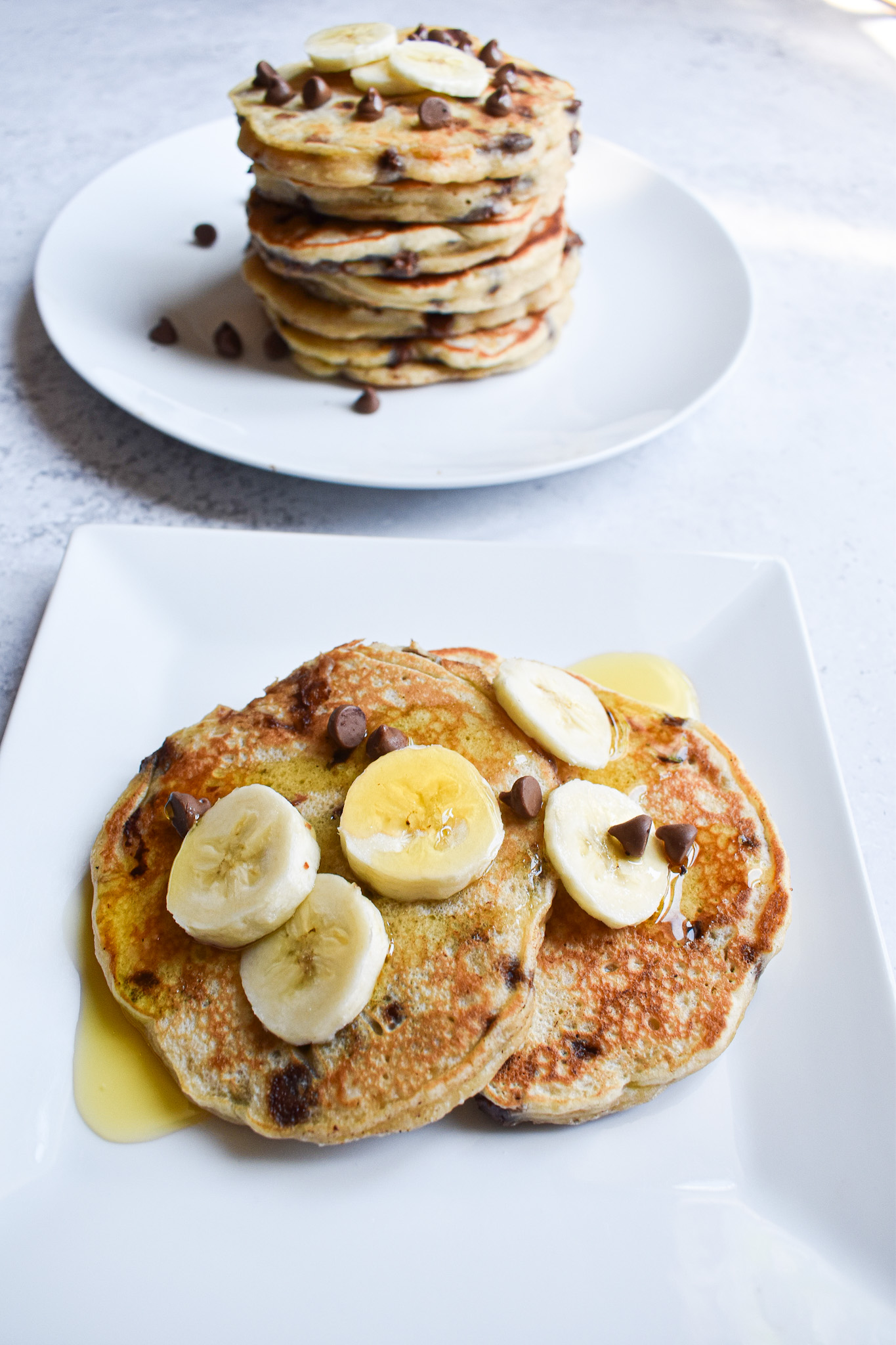 A white plate with 2 pancakes with syrup and chopped bananas. A stack of pancakes behind the front plate.